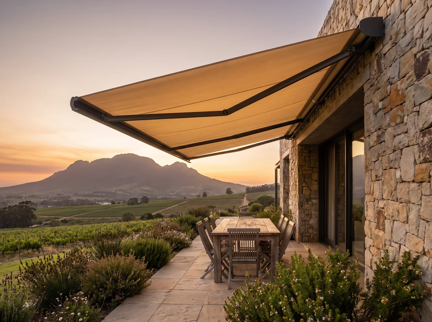 Retractable folding-arm awning extended over an outdoor dining table on a stone terrace in Paradyskloof, vineyard and mountain view at sunset